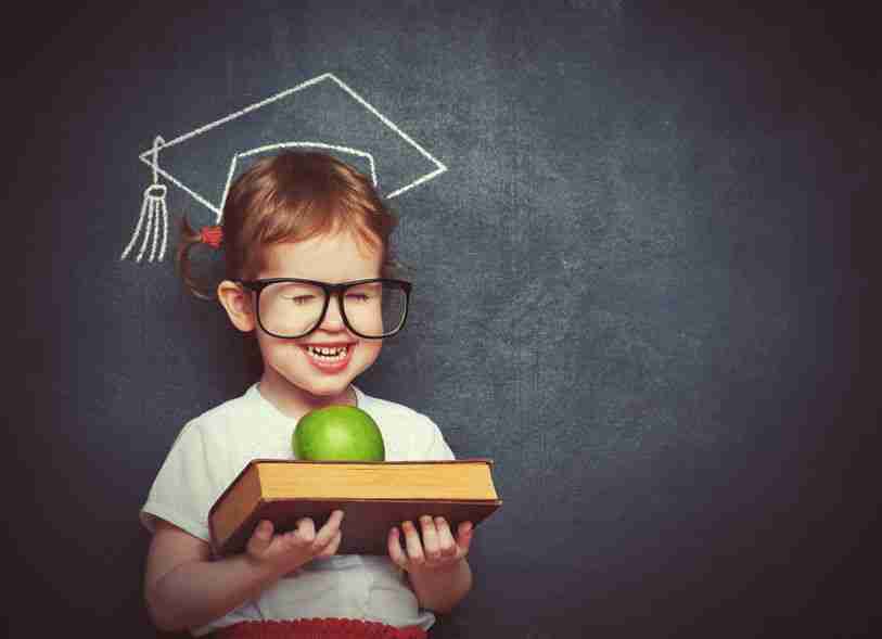 child wearing glasses holding a book with a green apple on top of it. They are standing in front of a chalkboard with an outlined graduation cap drawn in chalk.