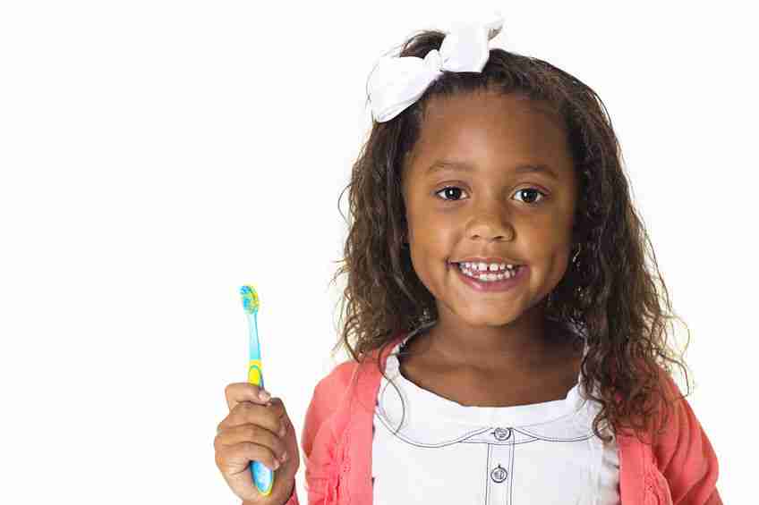 Little girl wearing a white bow and holding a toothbrush. Shes wearing a white shirt and a pink cardigan.