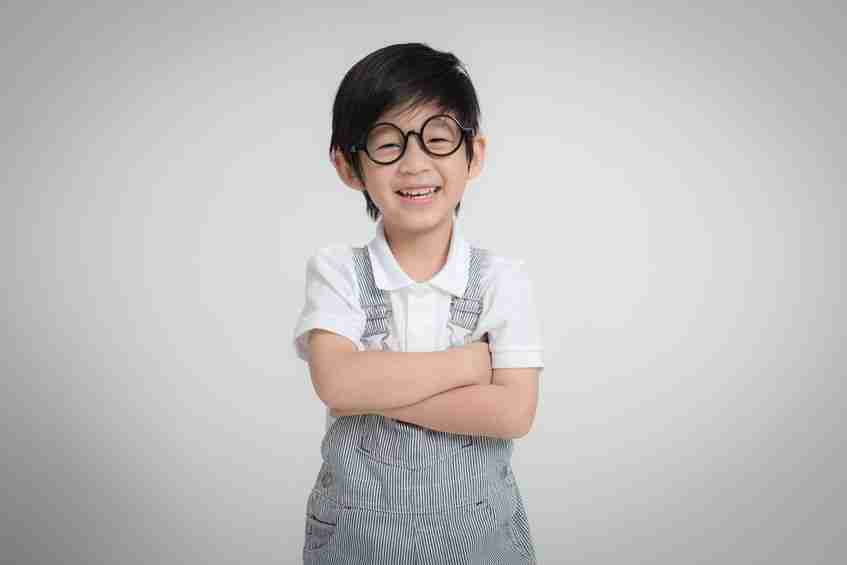 kid with arms crossed wearing glasses, gray overalls a a white collared shirt standing in front of a gray background