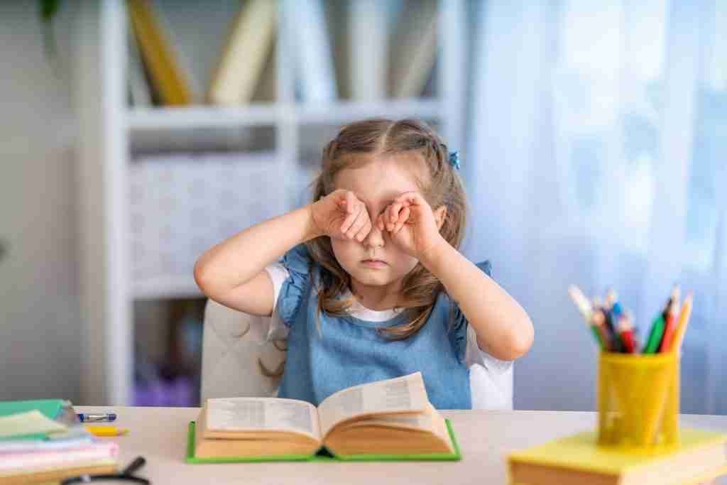 child is tired of learning. home schooling, homework. girl rubs his eyes from fatigue reading books and textbooks. little kid student sitting at table with books. vision problems